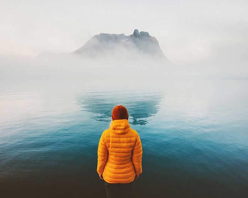 Woman thinking how do I find myself while contemplating water and rock visible through mist, wearing yellow jacket and red knit cap.